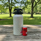 A large insulated water bottle covered in iridescent rhinestones stands on a wooden picnic table outdoors next to a red Coca-Cola can for scale. The background shows a blurred park with green grass and trees.